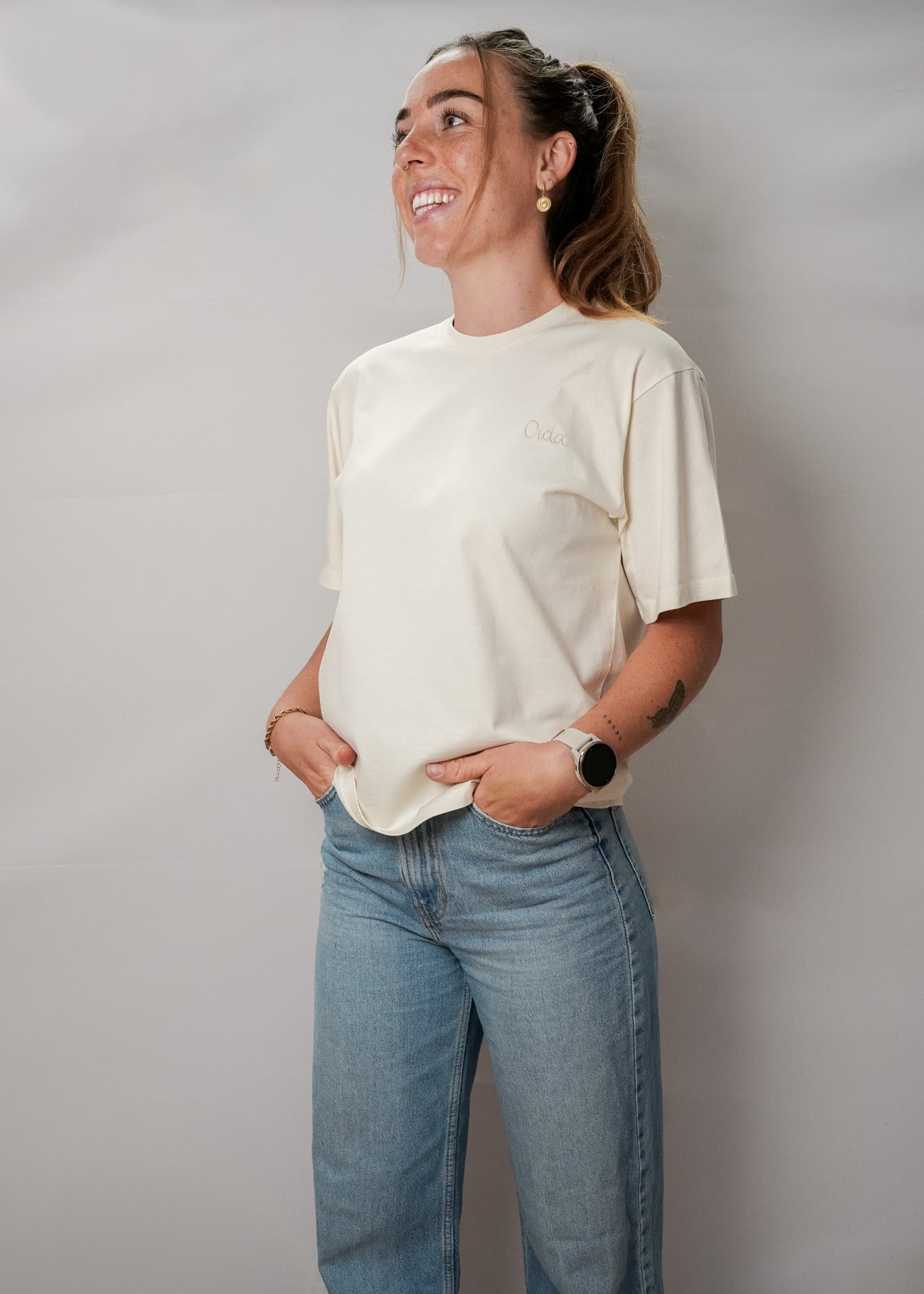Eine Frau mit langen, blonden Haaren, die zu einem hohen Pferdeschwanz gebunden sind, steht mit dem Rücken zur Kamera. Sie trägt ein cremefarbenes T-Shirt mit einem kleinen Stick des Worts "OIDA." Die Ärmel sind kurz, und das Shirt sitzt locker. Sie kombiniert es mit einer hellblauen Jeans. Der Hintergrund ist eine schlichte, graue Wand.