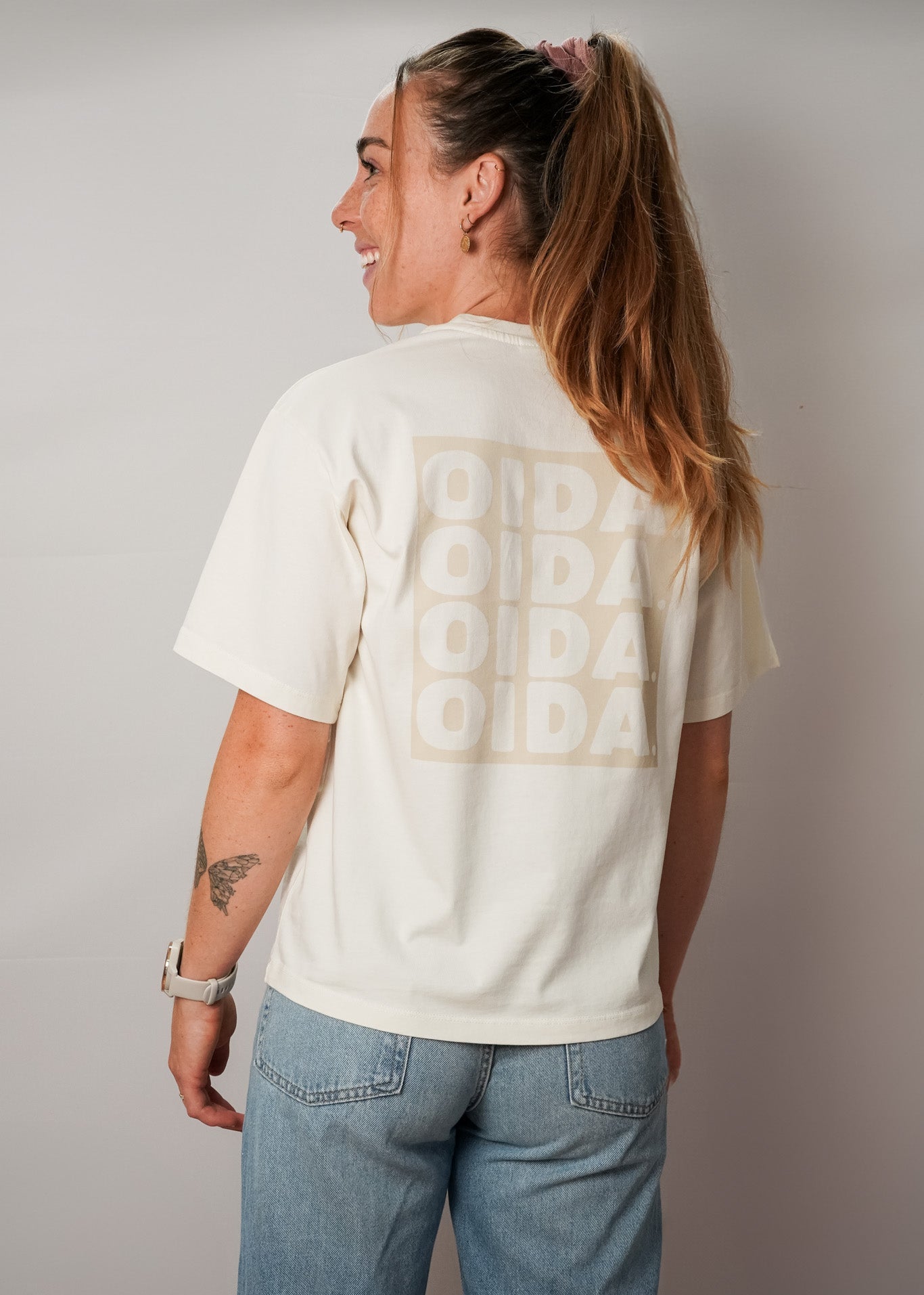 Eine Frau mit langen, blonden Haaren, die zu einem hohen Pferdeschwanz gebunden sind, steht mit dem Rücken zur Kamera. Sie trägt ein cremefarbenes T-Shirt mit einem großen, hellen Print auf dem Rücken, auf dem viermal das Wort "OIDA." in Blockbuchstaben steht. Die Ärmel sind kurz, und das Shirt sitzt locker. Sie kombiniert es mit einer hellblauen Jeans.