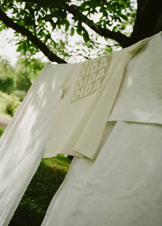 Alt-Text:  
Ein cremefarbenes T-Shirt mit dem aufgedruckten Wort "OIDA" in vier Reihen hängt auf einer Wäscheleine im Freien. Das T-Shirt ist über ein großes, weißes Tuch gelegt, das ebenfalls auf der Leine hängt. Im Hintergrund sind grüne Blätter eines Baumes zu sehen, durch die Sonnenlicht scheint.