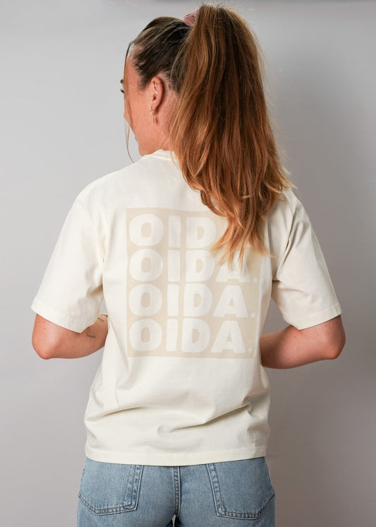 Eine Frau mit langen, blonden Haaren, die zu einem hohen Pferdeschwanz gebunden sind, steht mit dem Rücken zur Kamera. Sie trägt ein cremefarbenes T-Shirt mit einem großen, hellen Print auf dem Rücken, auf dem viermal das Wort "OIDA." in Blockbuchstaben steht. Die Ärmel sind kurz, und das Shirt sitzt locker. Sie kombiniert es mit einer hellblauen Jeans. Der Hintergrund ist eine schlichte, graue Wand.