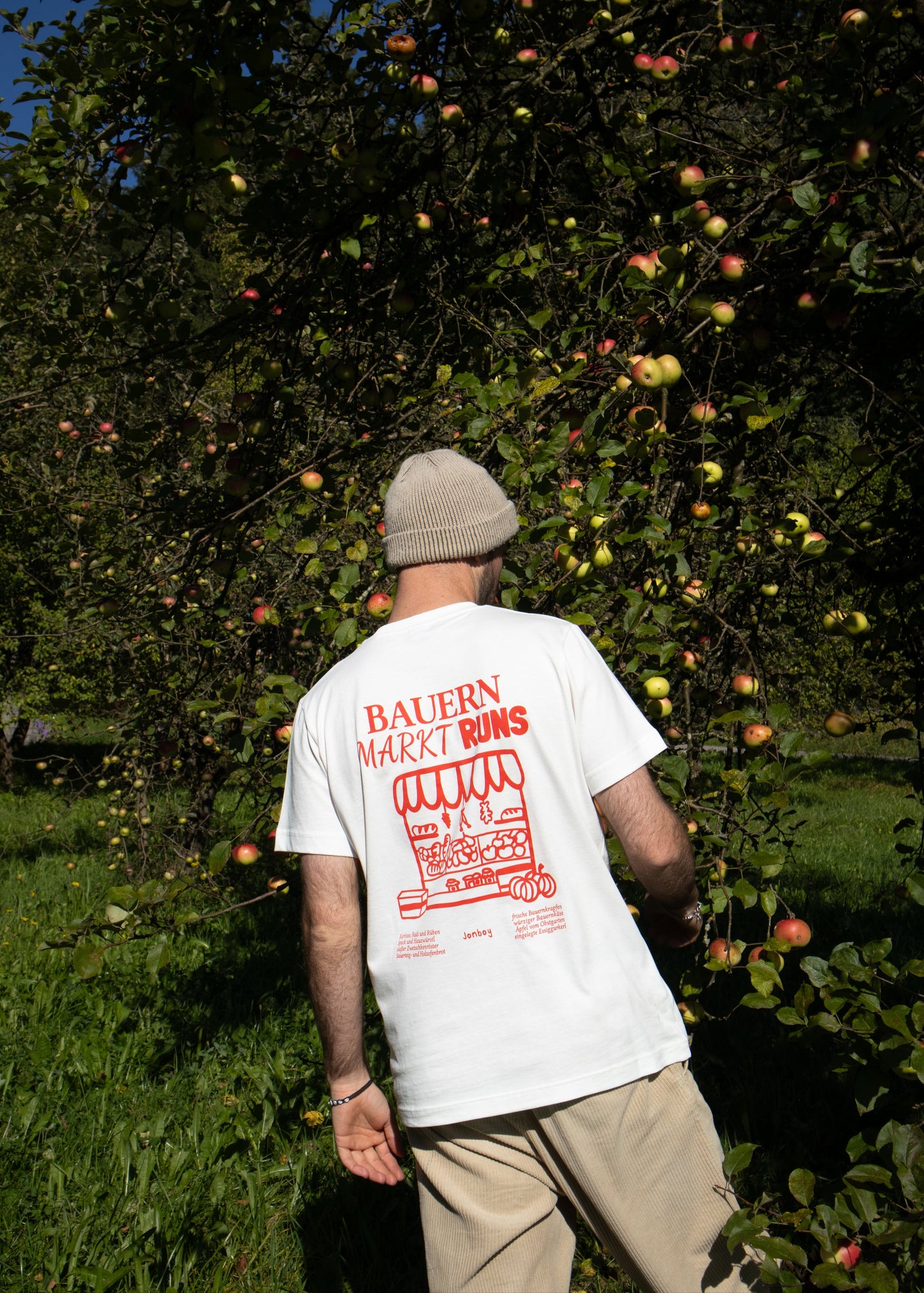 Mann steht mit dem Rücken vor einem Apfelbaum und trägt ein weißes T-Shirt mit rotem Bauernmarkt-Print.