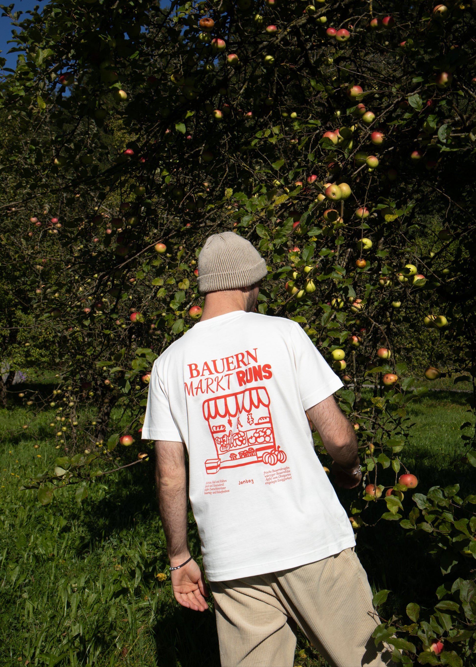 Mann steht mit dem Rücken vor einem Apfelbaum und trägt ein weißes T-Shirt mit rotem Bauernmarkt-Print.